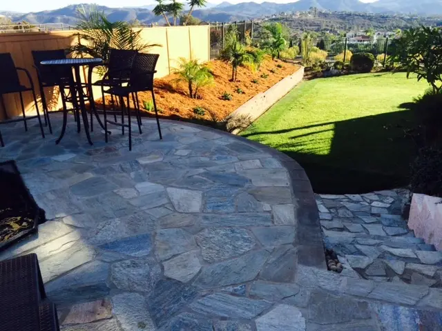 Stone patio with outdoor table and chairs, landscaped garden with palm plants, lawn, and distant mountains under a clear sky.