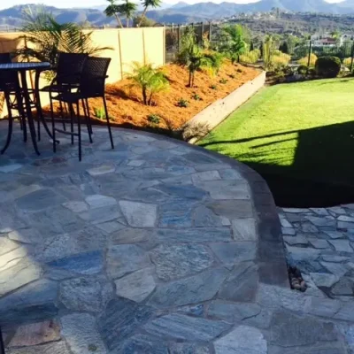 Stone patio with outdoor table and chairs, landscaped garden with palm plants, lawn, and distant mountains under a clear sky.